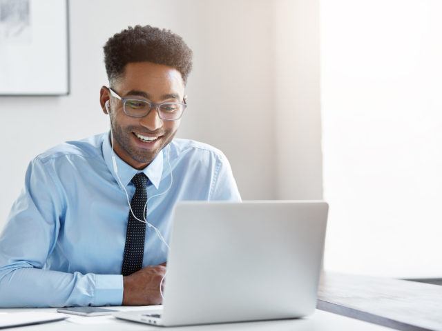 Indoor office shot of cheerful successful young African American manager with stubble sitting in front of open laptop wearing earphones while having video conference call with business partners