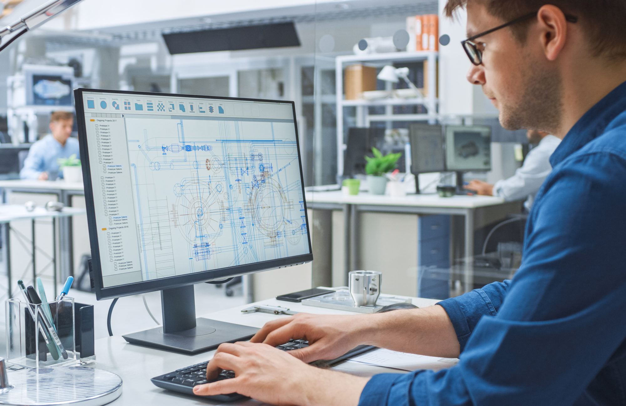 Over the Shoulder Shot of Engineer Working with CAD Software on Desktop Computer, Screen Shows Technical Draughts and Drawings. In the Background Engineering Facility Specialising on Industrial Design
