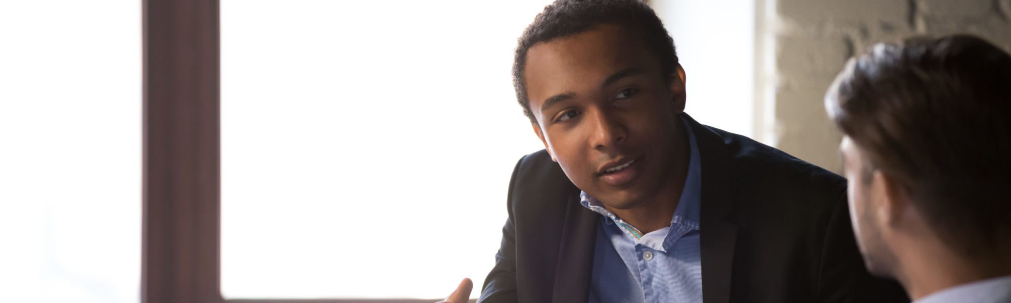 Focused African American male businessman talk with colleagues, explain business project sitting at table, diverse employees negotiate in office, discussing business issues or contract terms