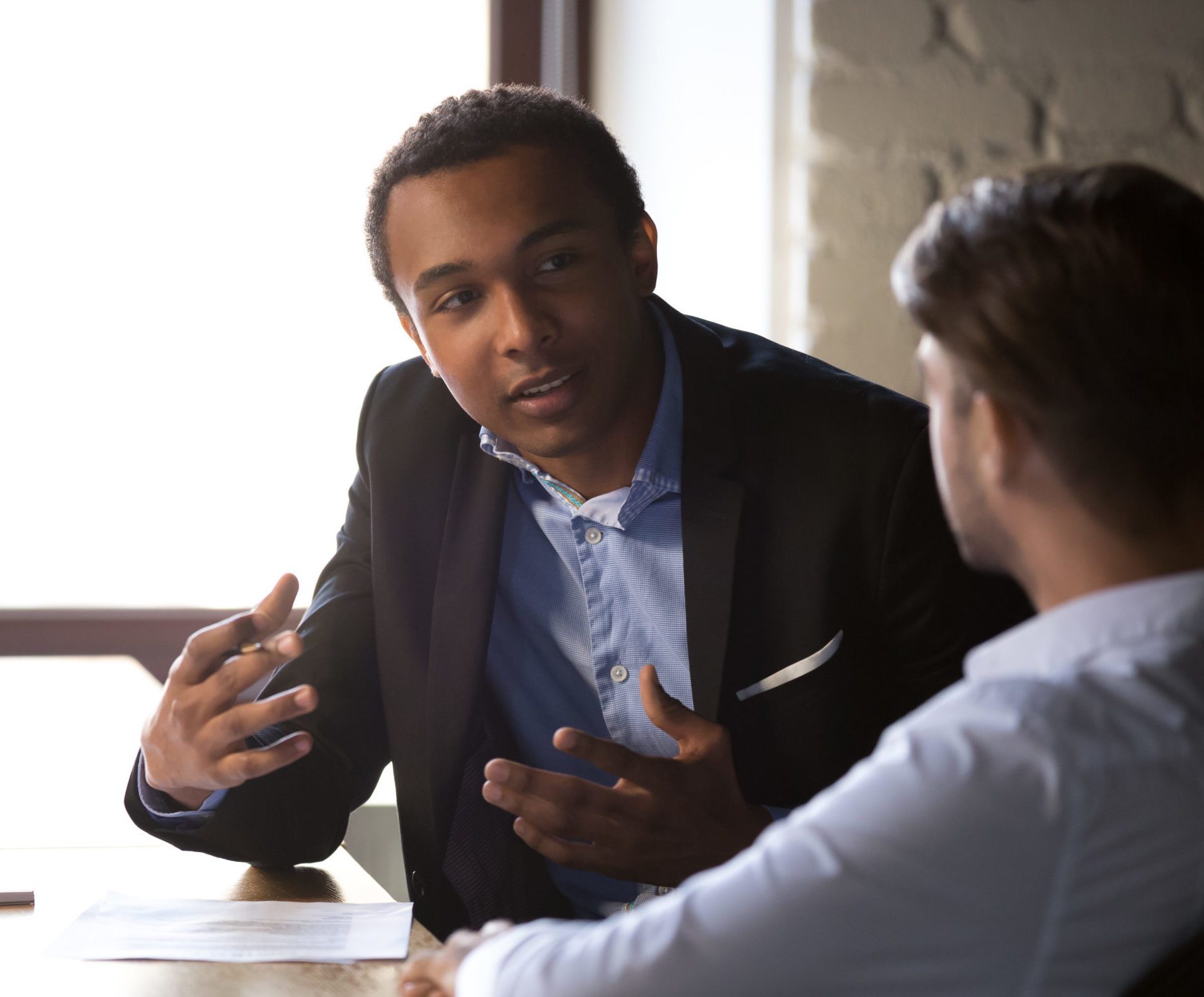 Focused African American male businessman talk with colleagues, explain business project sitting at table, diverse employees negotiate in office, discussing business issues or contract terms